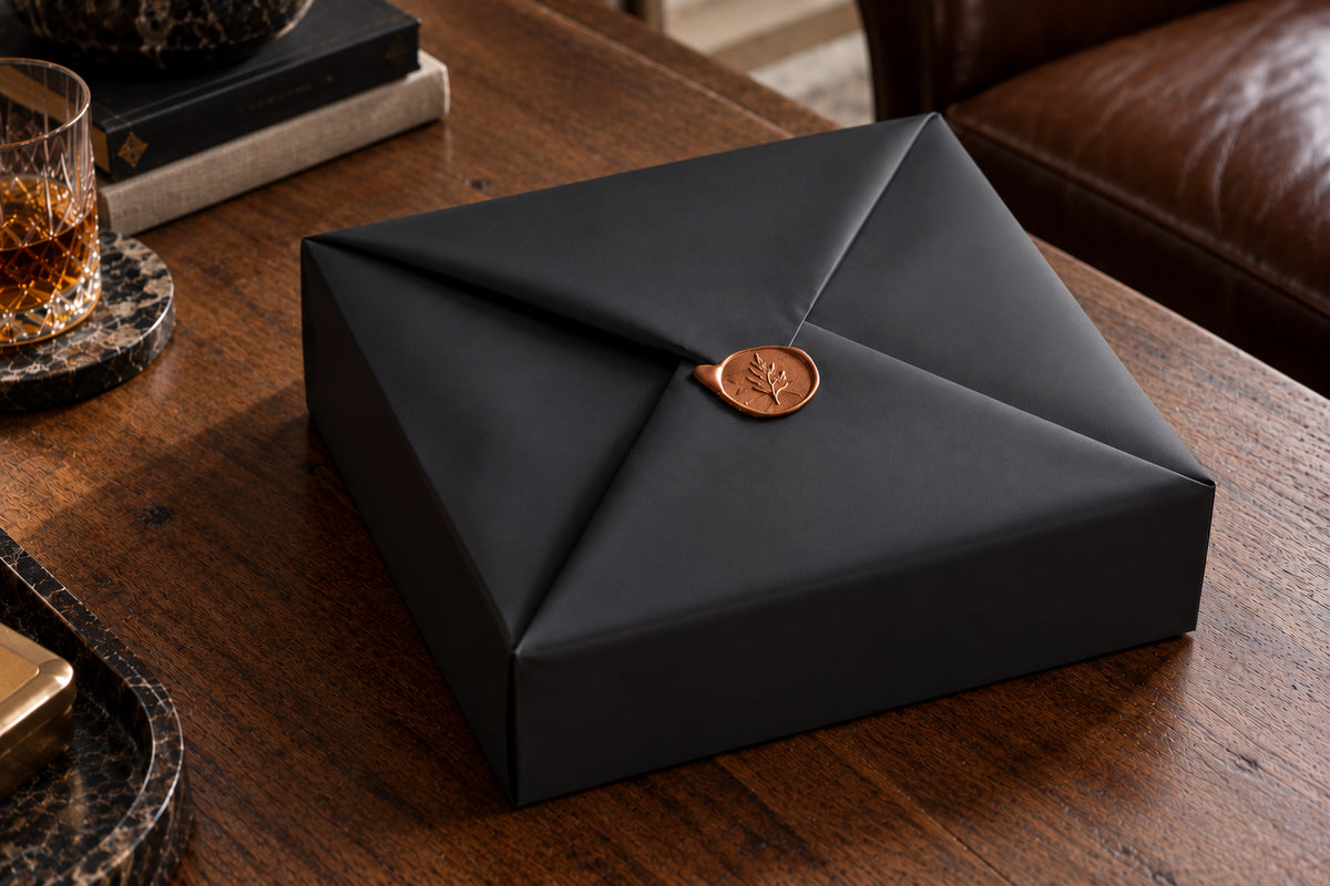Luxury matte-black gift packaging by Oak & Ember, featuring elegant envelope-style folds and a handcrafted bronze-colored wax seal, presented on a dark wooden table.
